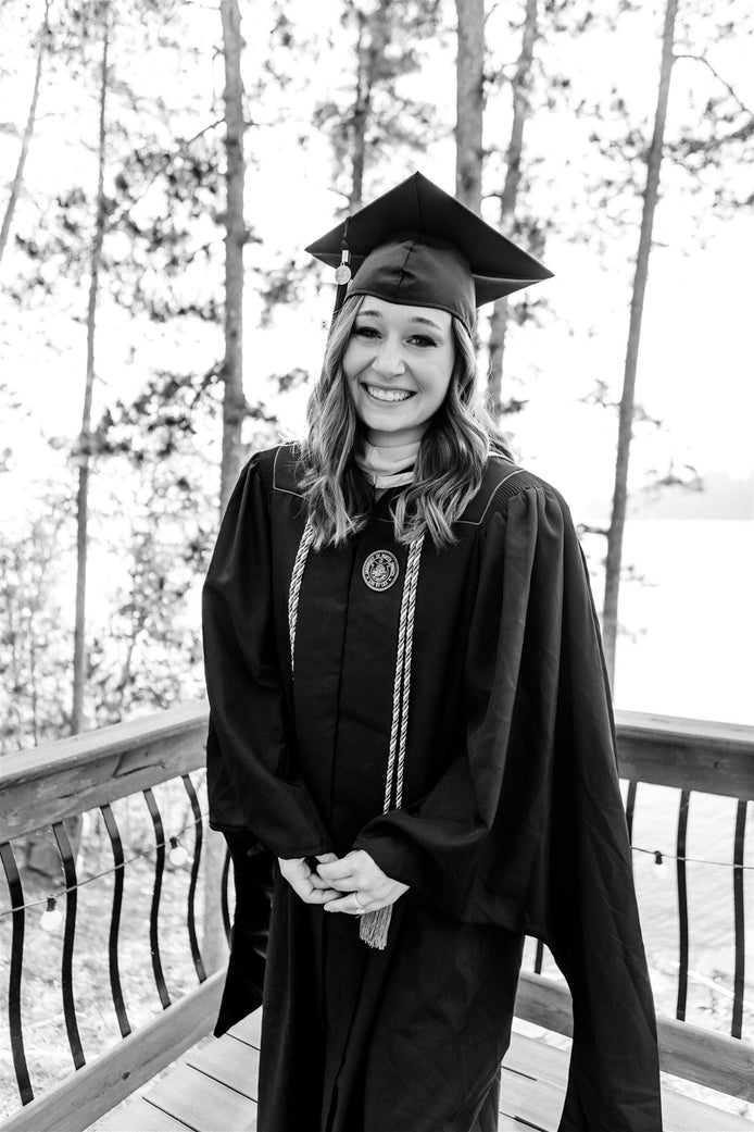 Smiling woman in graduation cap and gown standing on a wooden deck with trees in the background.
